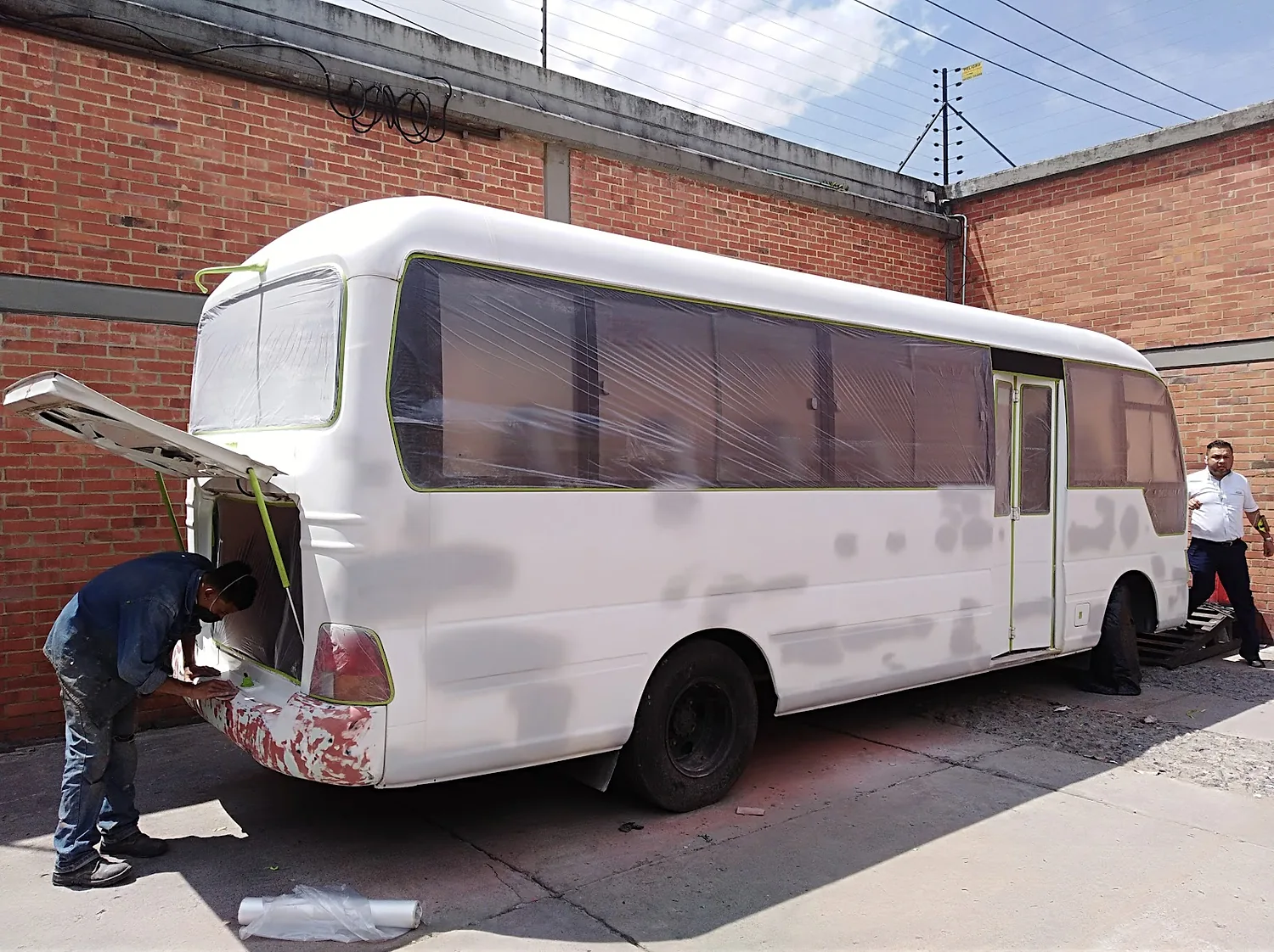 Un operario de Lottus Express trabaja el panel trasero de un microbús enmascarado con plástico y cinta, listo para pintar, en el patio de la Calle 163